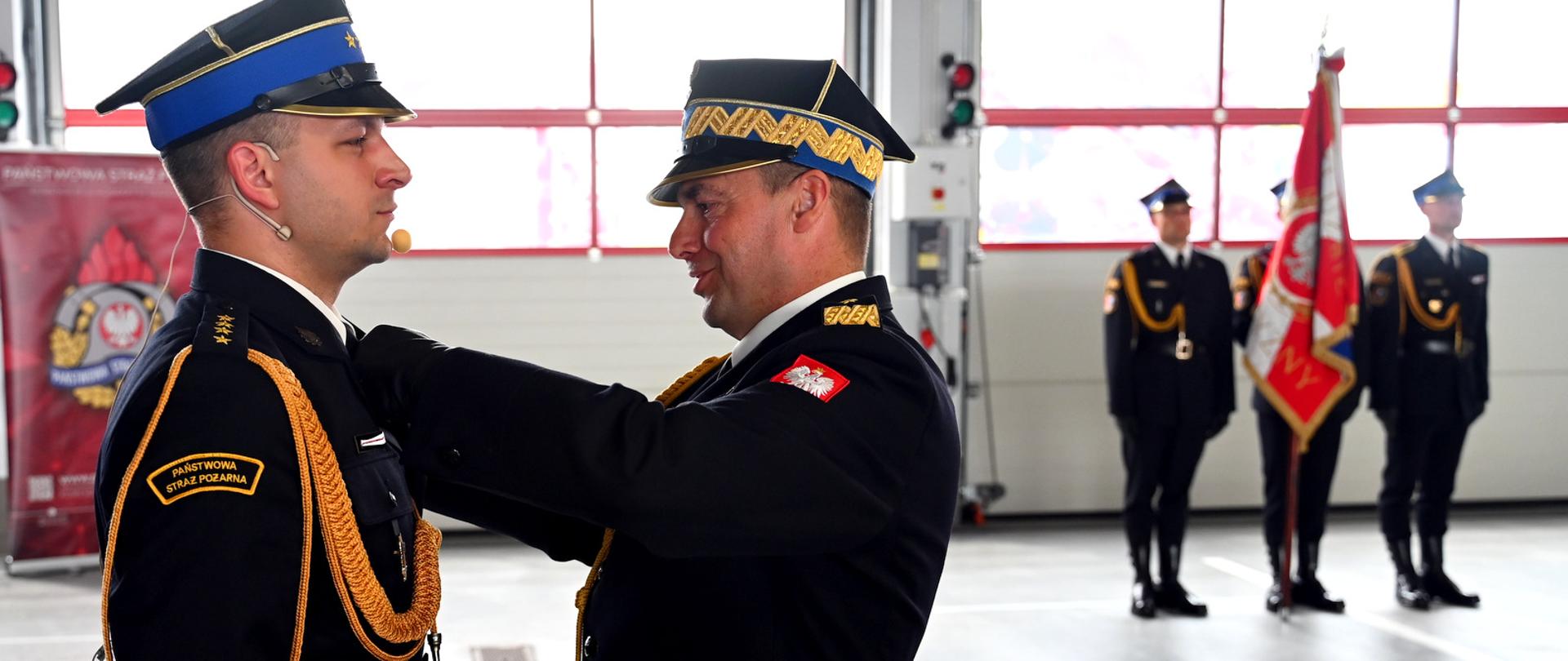 Panorama hali JRG nr 3 w Rzeszowie. Nadbryg. Tomasz Baran przypina odznaczenie oficerowi z mikrofonem nagłownym. W tle, przed wielkimi oknami garażowymi, stoi na baczność poczet sztandarowy oraz inni strażacy.
