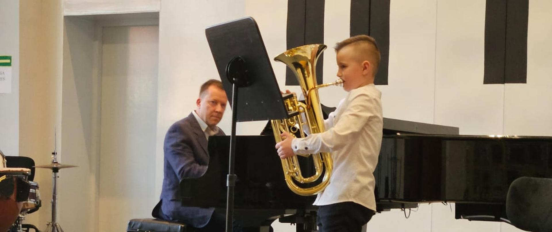 Chłopiec gra na sakshornie, mężczyzna akompaniuje na fortepianie.