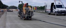 Na zdjęciu widać świeżo ułożoną nawierzchnię betonową, na niej maszyna wykonująca szczeliny dylatacyjne, obsługiwana przez robotnika. W dalszej perspektywie po lewej stronie widać maszyny drogowe, po prawej stojący bus, a w oddali las. 