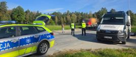 Na zdjęciu widać wspólne działania kontrolne Inspekcji Transportu Drogowego i Policji. Na pierwszym planie znajduje się oznakowany samochód Policji oraz samochód Inspekcji Transportu Drogowego. W tle inspektor ITD i policjant prowadzą kontrolę pojazdu ciężarowego z naczepą. Na miejscu znajduje się również kierowca. Kontrola odbywa się na placu przy drodze, w otoczeniu terenów leśnych.