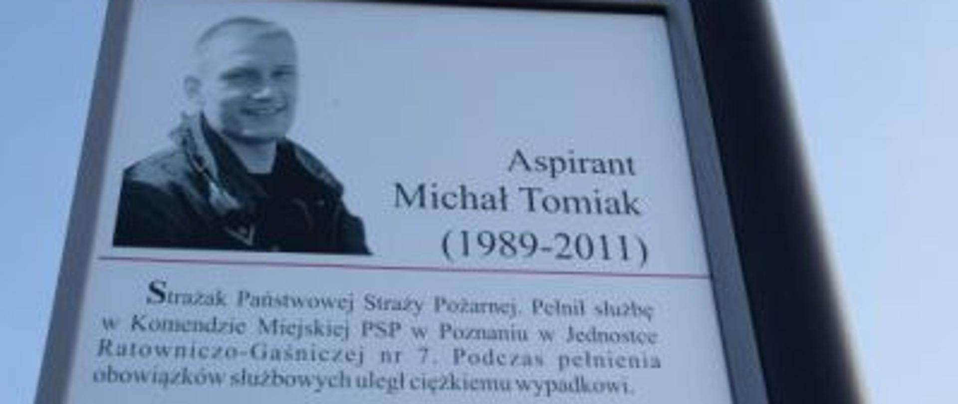 Metalowy słupek zamontowany przy ulicy, na którym widnieje tablica ze zdjęciem i wpisem upamiętniającym Michała Tomiaka