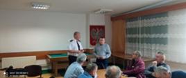 Zdjęcie wykonane na świetlicy komendy. W tle na ścianie widać godło Polski. Ściany jasne. Na środku zdjęcia strażak w białej koszuli a obok niego osoba ubrana w niebieską koszulę. Na pierwszym planie siedzący inni uczestnicy spotkania.