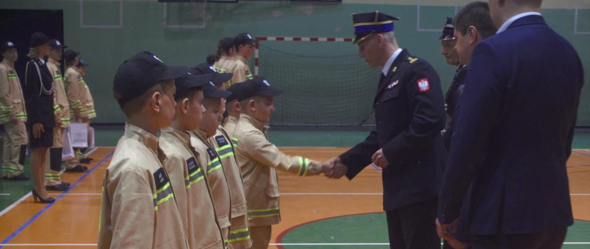 W pomieszczeniu, w szeregu stoi pięcioro dzieci z Drużyny Pożarniczej, ubrani w mundurki koloru piaskowego. Przed nimi zaproszeni goście, Komendant Powiatowy st. bryg. Janusz Bula w umundurowaniu wyjściowym z uściskiem z dłoni z jednym z dzieci.