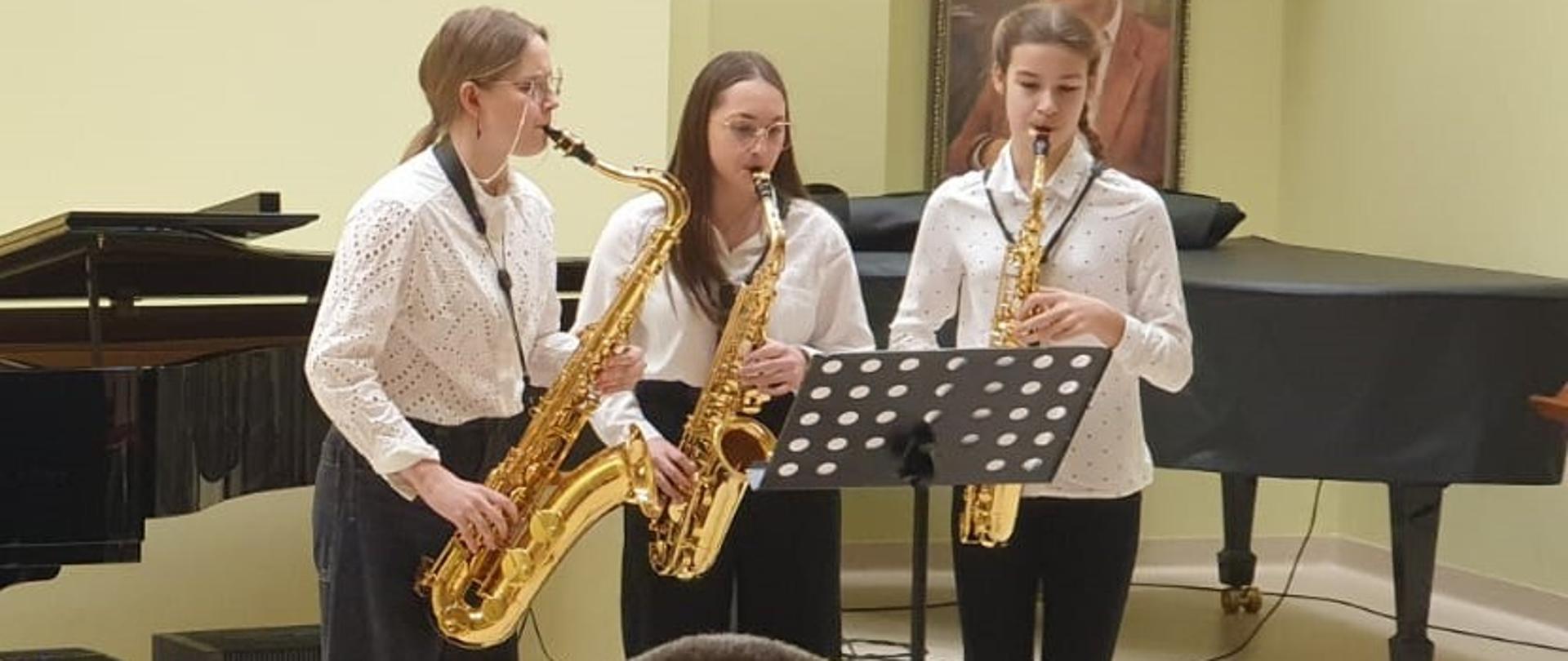 **Opis alternatywny:**
Trzy młode saksofonistki w białych koszulach i czarnych spodniach występują na scenie podczas XXXIX Konkursu Zespołów Kameralnych Szkół Muzycznych I i II stopnia. Stoją przed pulpitem z nutami, grając na saksofonach altowych. W tle fortepian. W sali dominuje jasna kolorystyka z pastelowozielonymi ścianami.
