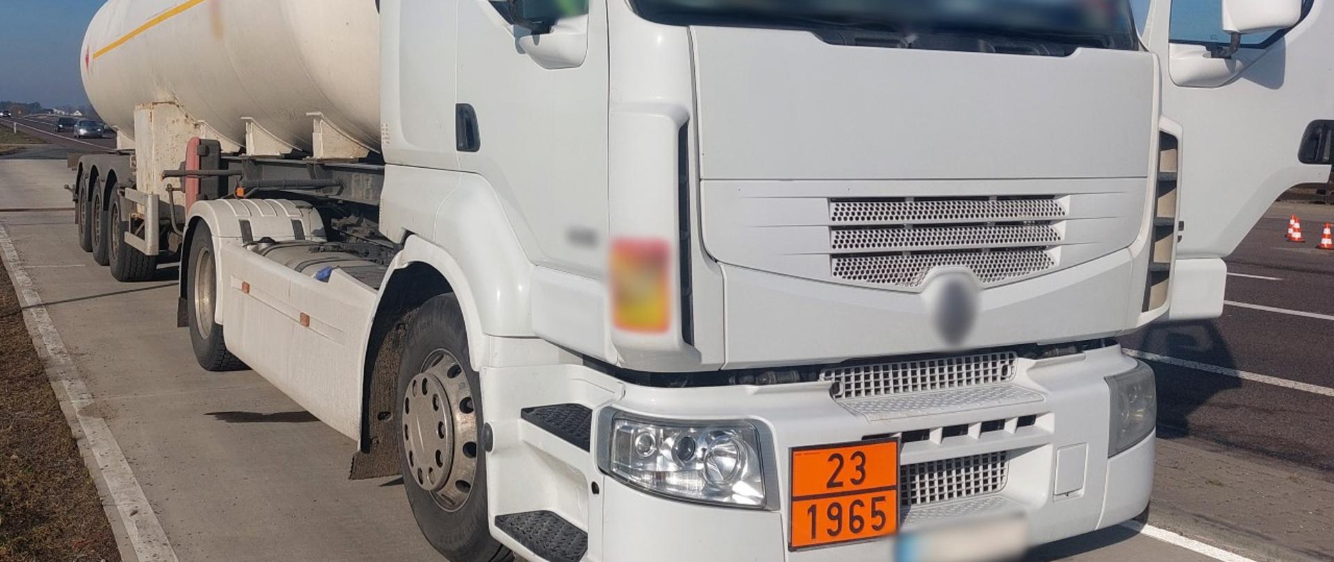Na pierwszym planie: przód ciągnika siodłowego z podpiętą naczepą-cysterną. W tle: samochody przejeżdżające krajową „dwunastką” obok punktu kontrolnego. 
