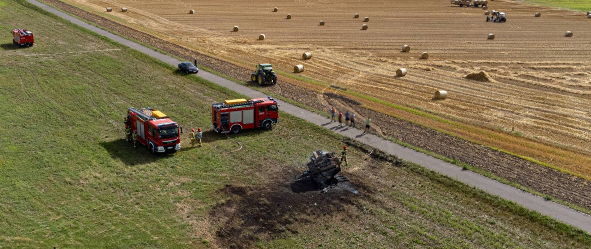 Zdjęcie z lotu ptaka przedstawia spaloną balotnicę na polu. W tle znajdują się samochody pożarnicze. 