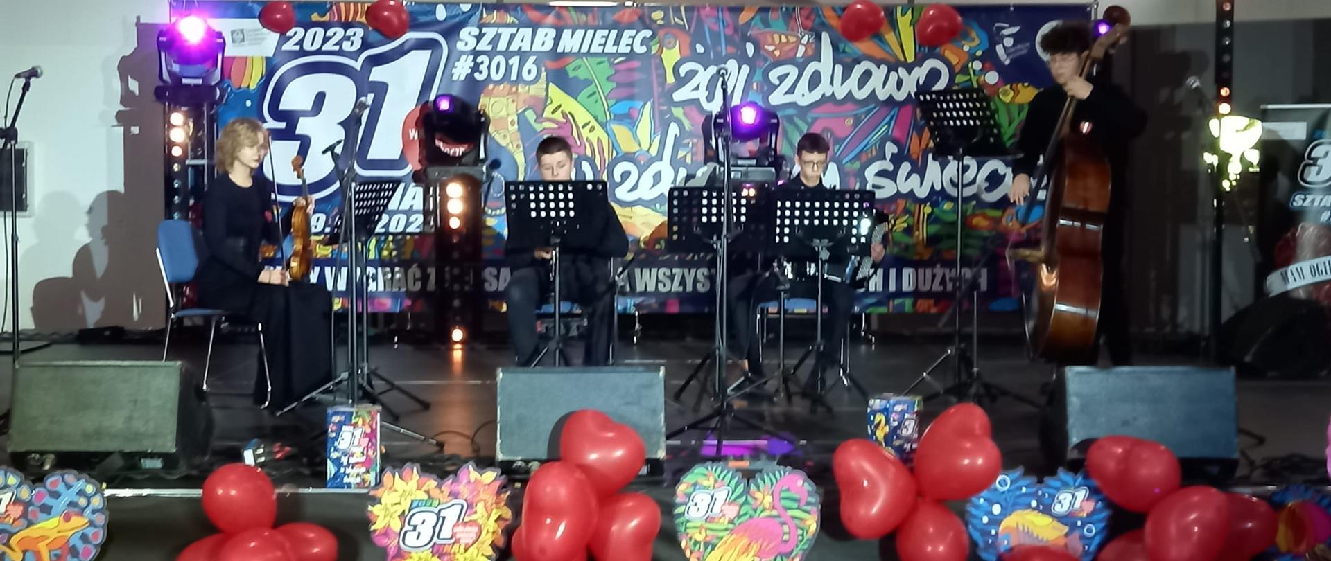 zdjęcie przedstawia na pierwszym planie rozsypane czerwone balony w kształcie serc, dalej czarną scenę, a na niej zespół kameralny złożony ze skrzypiec, klarnetu, akordeonu i kontrabasu, w tle widać duży napis 31. finał WOŚP oraz numer mieleckiego sztabu #3016