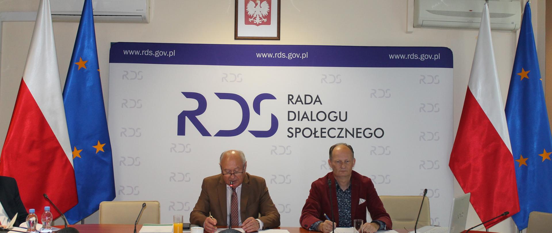 Na zdjęciu widnieje stół prezydialny za którym siedzą: Pan Jan Klimek, Przewodniczący Zespołu oraz Pan Norbert Pruszanowski, Związek Rzemiosła Polskiego. W tle znajduje się baner z napisem Rada Dialogu Społecznego. Po jego obu stronach znajdują się po dwie flagi: Polski i Unii Europejskiej. Nad banerem wisi Godło Polski. 