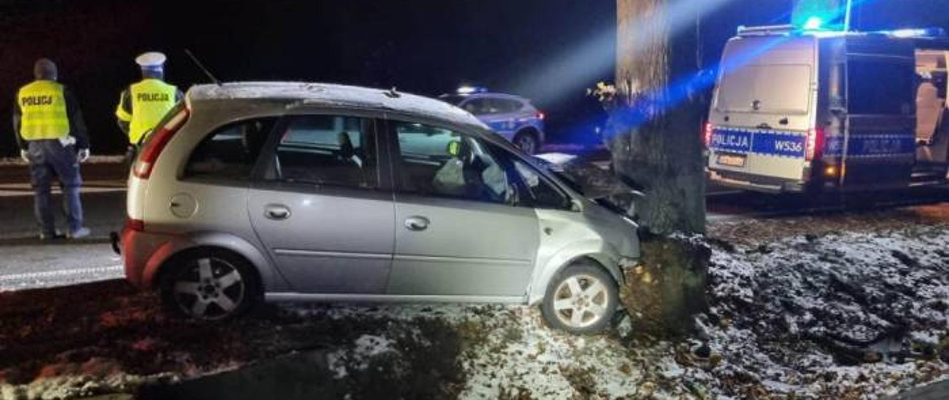 Samochód osobowy z mocno uszkodzonym przodem oparty o drzewo, dwa radiowozy i dwóch policjantów.