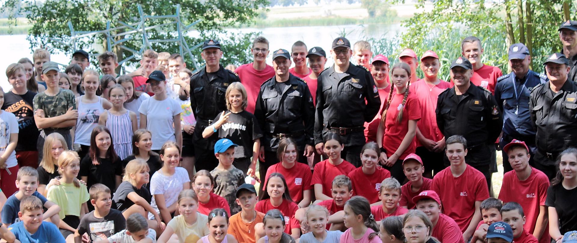 Zdjęcie grupowe dzieci, strażaków i wychowawców obozu harcerskiego - zbliżenie na centralne postacie trzech stojących wśród dzieci strażaków 