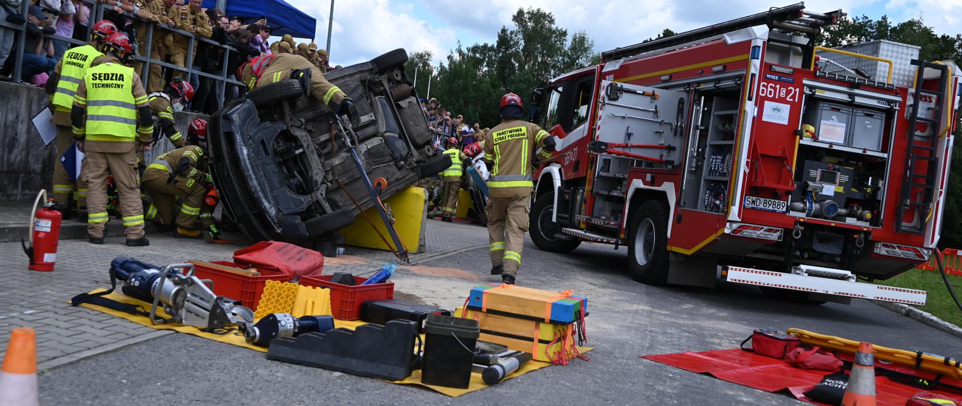 Pokaz ratownictwa technicznego straży pożarnej na terenie otwartym. Po lewej stronie widać ciemny samochód osobowy przewrócony na bok, przy którym pracują strażacy. Po prawej stronie stoi duży, czerwony wóz strażacki z otwartymi skrytkami sprzętowymi. Na pierwszym planie, na żółtej macie, ułożone są specjalistyczne narzędzia hydrauliczne. W tle na podwyższeniu zgromadzona jest publiczność obserwująca akcję.