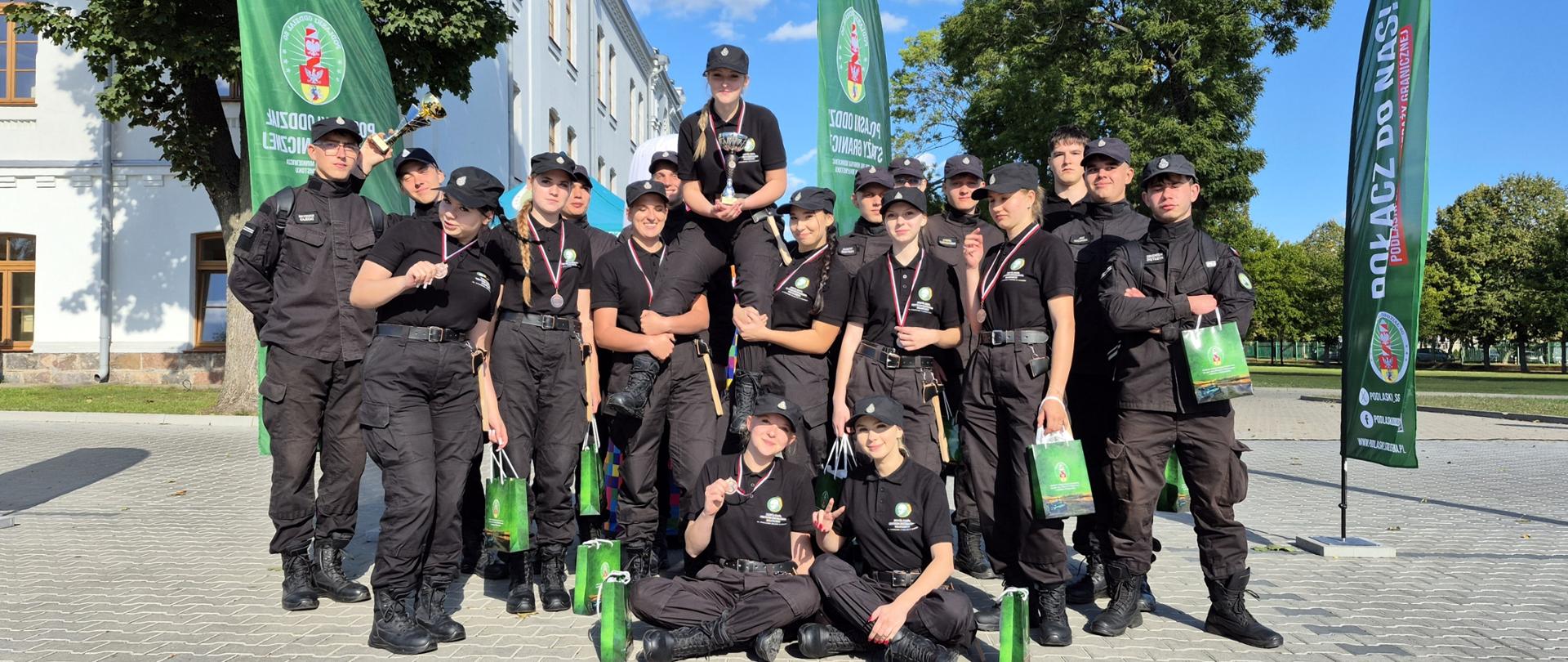 Grupa uczniów w czarnych mundurach, dowódcy drużyn - chłopak i dziewczyna - trzymają puchary, dziewczęta mają medale