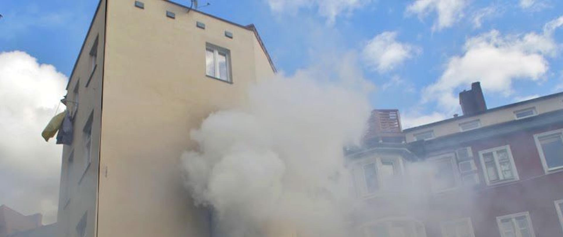Zdjęcie przedstawie kłęby dymu wydostające się z palącego mieszkania kamienicy na pierwszym piętrze. Przy budynku strażacy sprawiający drabinę.