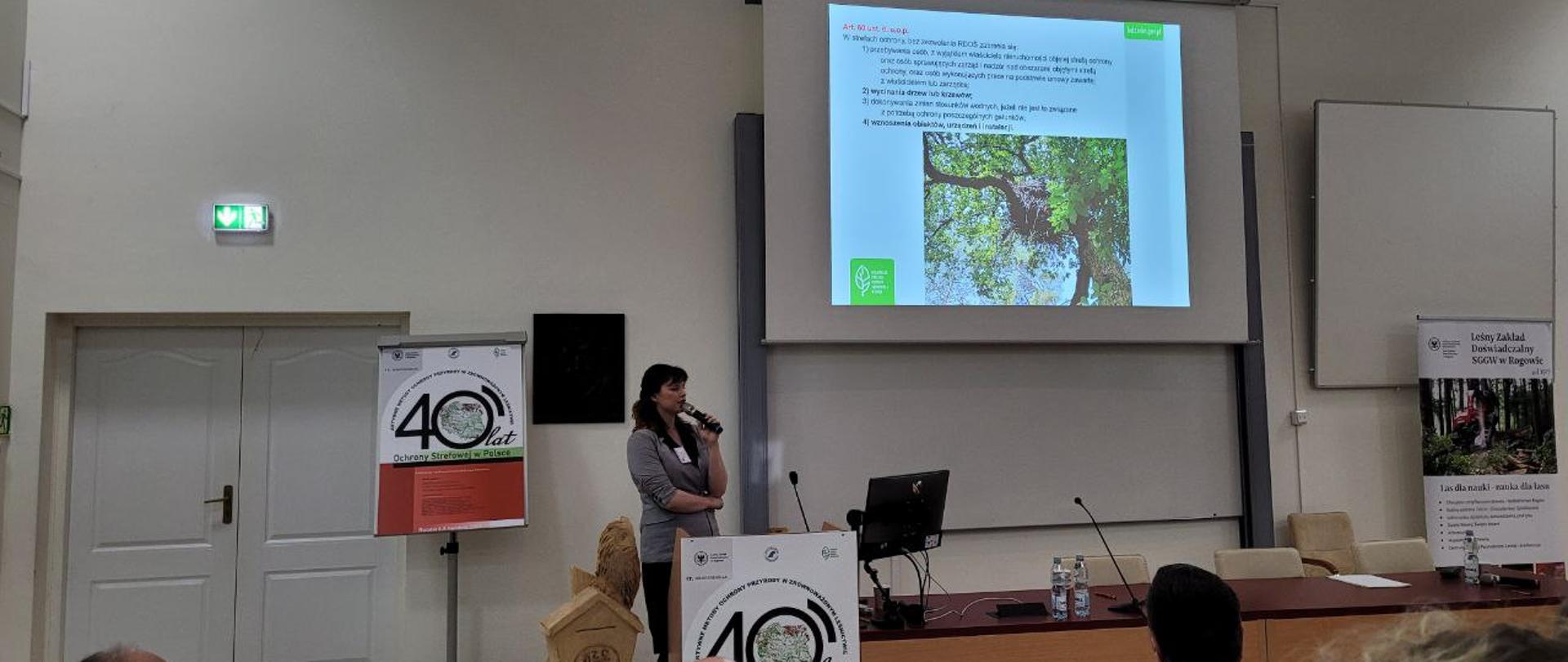 Kobieta stoi z mikrofonem, za nią ekran na którym widać zdjęcia drzewa i tekst. 