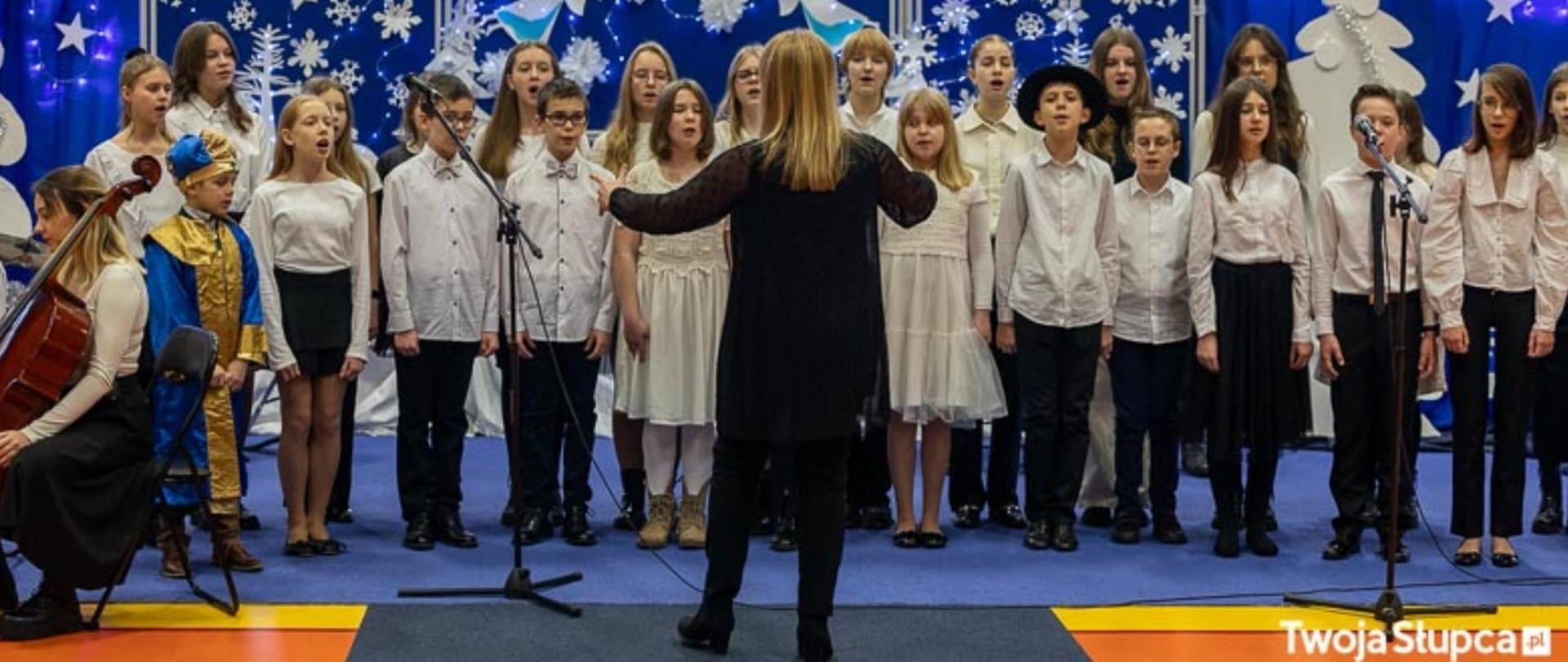 zdjęcie chóru i dyrygenta podczas występu na sali gimnastycznej na tle dekoracji świątecznych