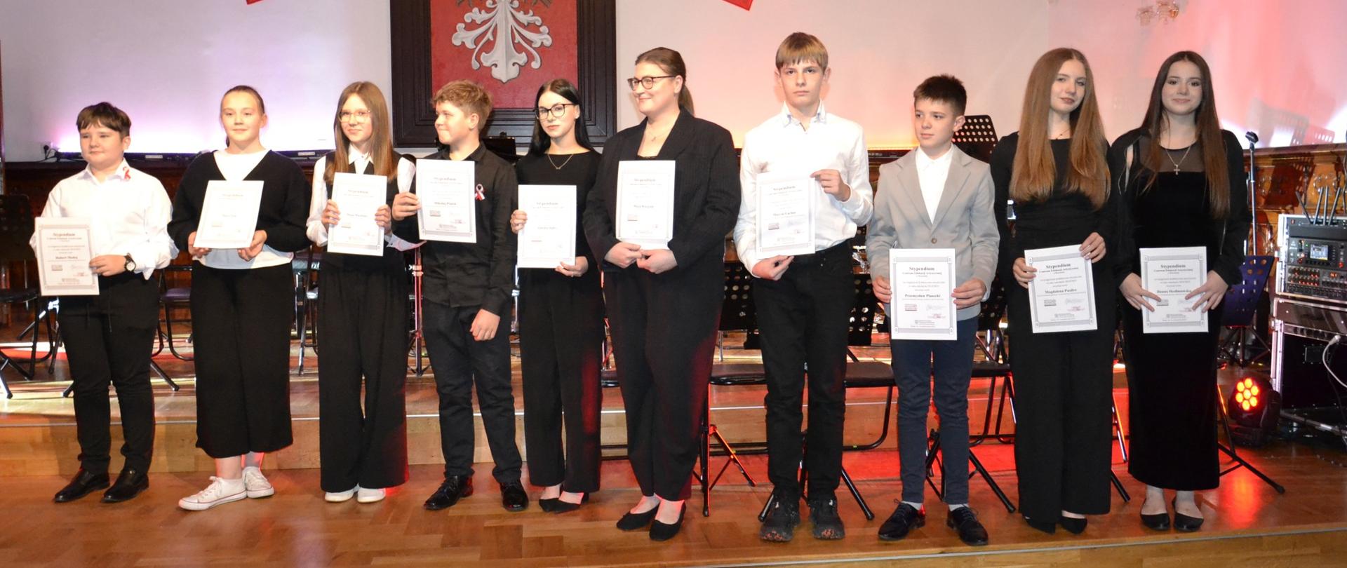 Grupa uczniów stoi na scenie z dyplomami w dłoniach podczas uroczystego wręczenia stypendiów Dyrektora Centrum Edukacji Artystycznej w trakcie Koncertu Patriotycznego.