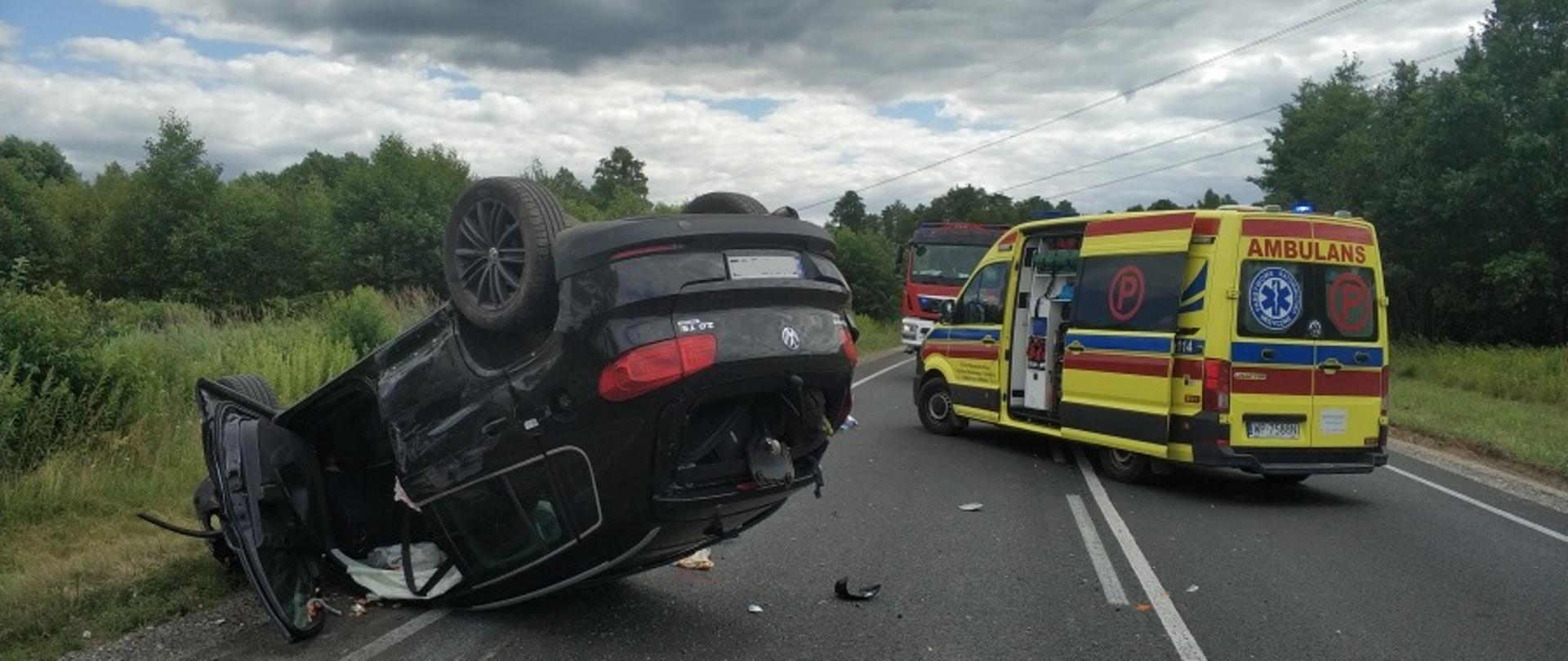 Na zdjęciu widoczny rozbity samochód osobowy marki Volkswagen koloru czarnego leżący na jezdni na dachu. Widoczne uszkodzenia karoserii oraz wystrzelone poduszki i kurtyny powietrzne. Na drugim planie żółty ambulans i samochód straży pożarnej.