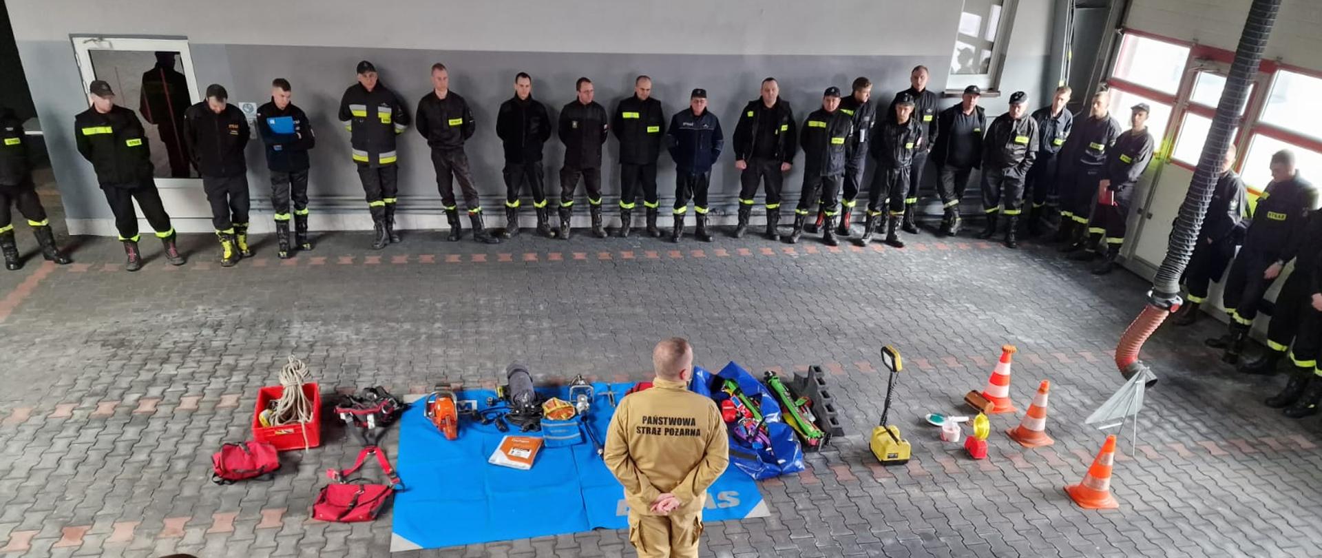 Na zdjęciu druhowie Ochotniczych Straży Pożarnych w umundurowaniu koszarowym ustawieni wokół stanowiska do instruktażu oraz funkcjonariusze Państwowej Straży Pożarnej prowadzący szkolenie z metodyki przeprowadzania instruktażu stanowiskowego z zakresu bezpieczeństwa i higieny pracy.