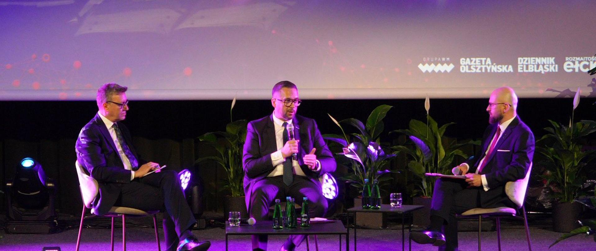 W zaciemnionej sali lekko oświetlonej na fioletowo na podwyższeniu na krzesłach siedzi trzech mężczyzn w garniturach, pośrodku minister Kulasek mówi do trzymanego w ręku mikrofonu.