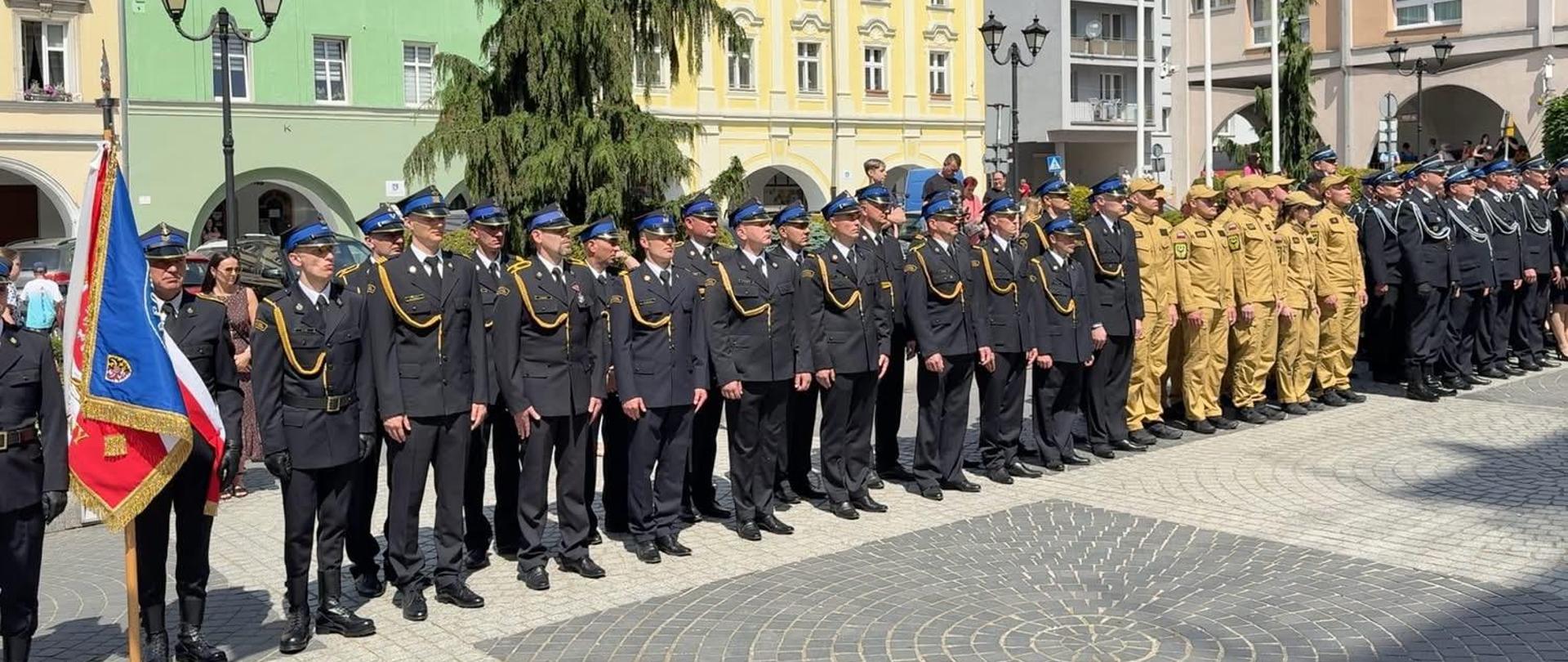 Obraz przedstawia strażaków Państwowej i Ochotniczych Straży Pożarnych stojących w dwuszeregu na płycie rynku w Jaworze. Strażacy w mundurach wyjściowych i służbowych. Po lewej stronie widoczny sztandar jaworskiej komendy. Pora dzienna