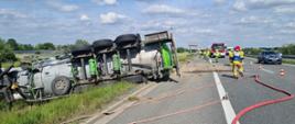 Zdjęcie zrobione zostało na autostradzie. Po prawej stronie widać jadące lewym pasem ruchu samochody. Następnie można zauważyć leżącą na jezdni linie wężową służącą do zabezpieczenia na wypadek ewentualnego pożaru. W oddali z prawej strony widać pojazd straży pożarnej oraz strażaków wraz z kierującym działaniem ratowniczym, który ma ubraną biało-niebieską kamizelkę. Po prawej stronie widać leżącą na boku cysternę. Widać jej podwozie wraz z uszkodzeniami. Zdjęcie wykonane zostało w słoneczny dzień.