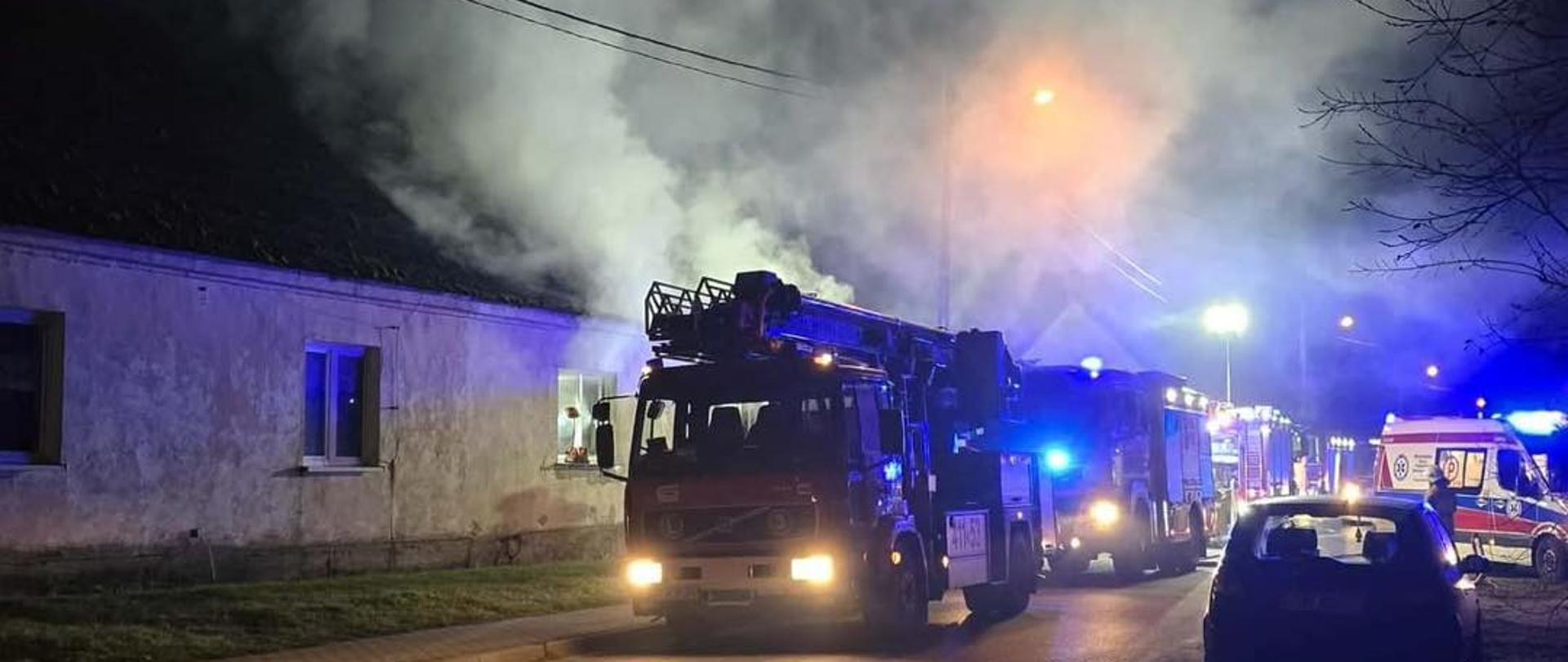 Nocna akcja gaśnicza przy długim, jasnym budynku mieszkalnym. Z dachu unosi się gęsty biały dym. Na ulicy stoi wóz strażacki z włączonymi światłami, a za nim kolejne pojazdy służb ratunkowych, w tym karetka. Okolica oświetlona jest niebieskim i pomarańczowym światłem sygnałów.