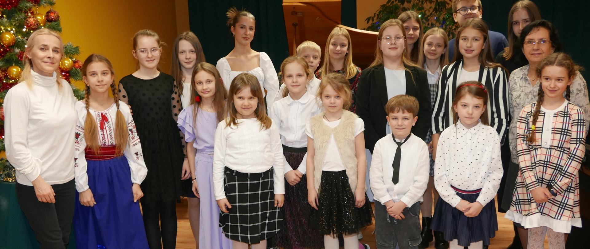 Zdjęcie grupowe uczniów klasy fortepianu. Z przodu stoją najmłodsi uczniowie, z tyłu najstarsi. z prawej strony za uczennicą w warkoczach i sukience w kratkę stoi nauczycielka w okularach z czarnymi włosami. po lewej stronie stoi druga nauczycielka o blond włosach w białym golfie i ciemnych spodniach