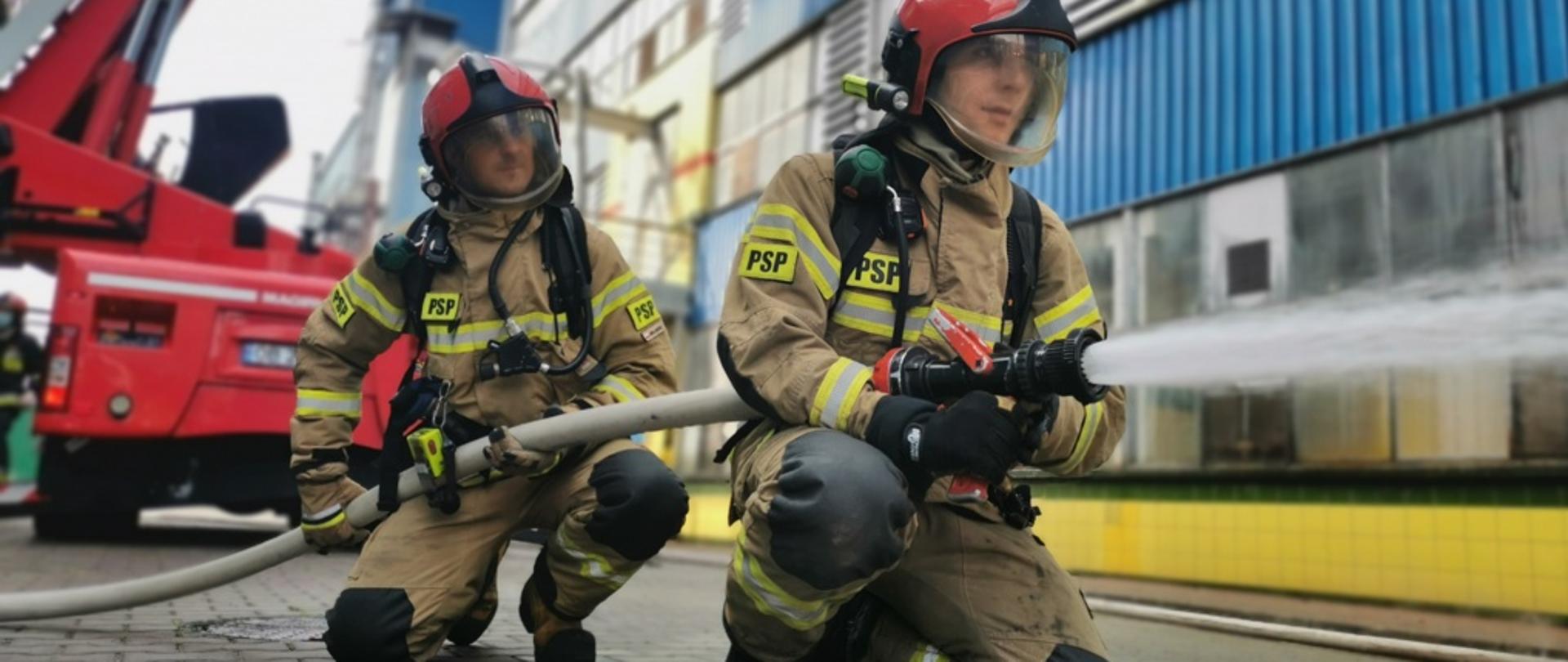 Dwóch strażaków podczas gaszenia pożaru, jeden trzyma prądownicę, drugi podtrzymuje wąż.