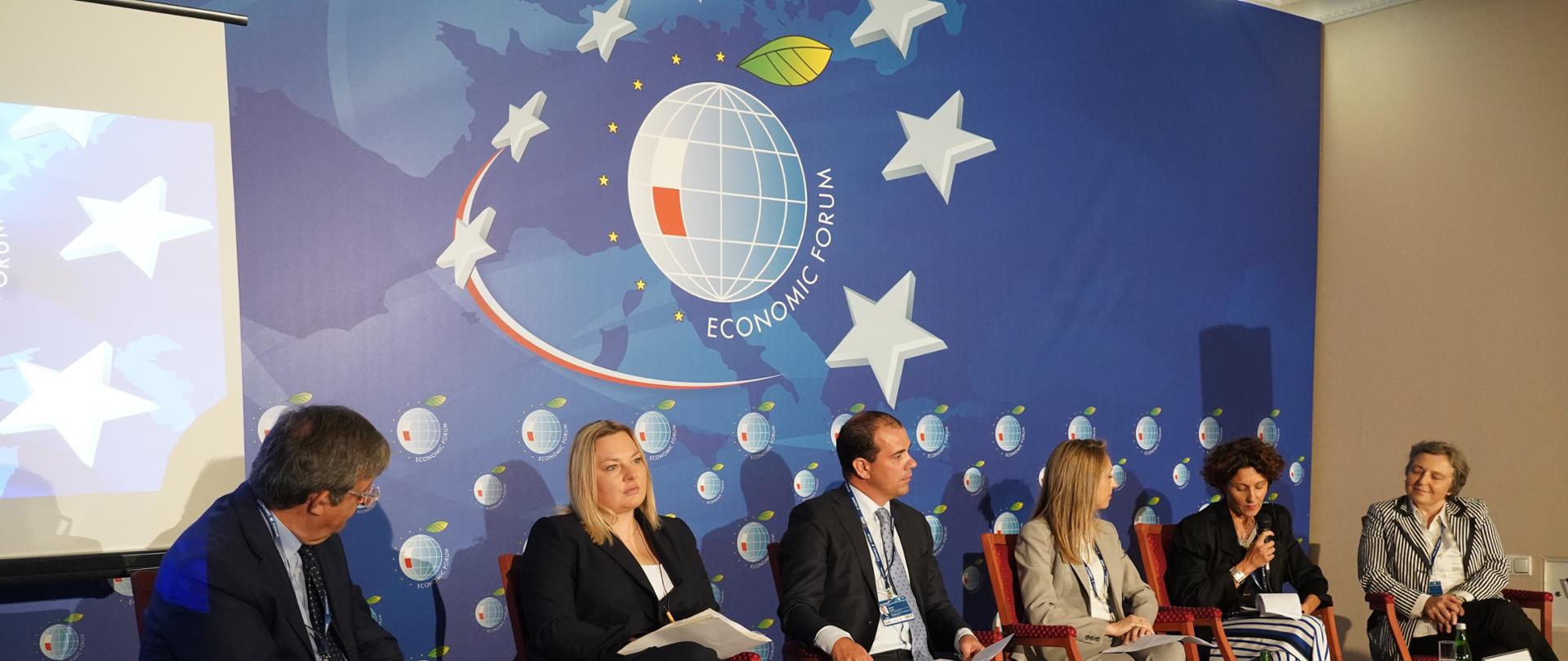 Wiceminister funduszy i polityki regionalnej Ewelina Owczarska wraz z pozostałymi panelistami na Forum Ekonomicznym w Karpaczu, siedzą za stolikami w tle logo organizatora kongresu