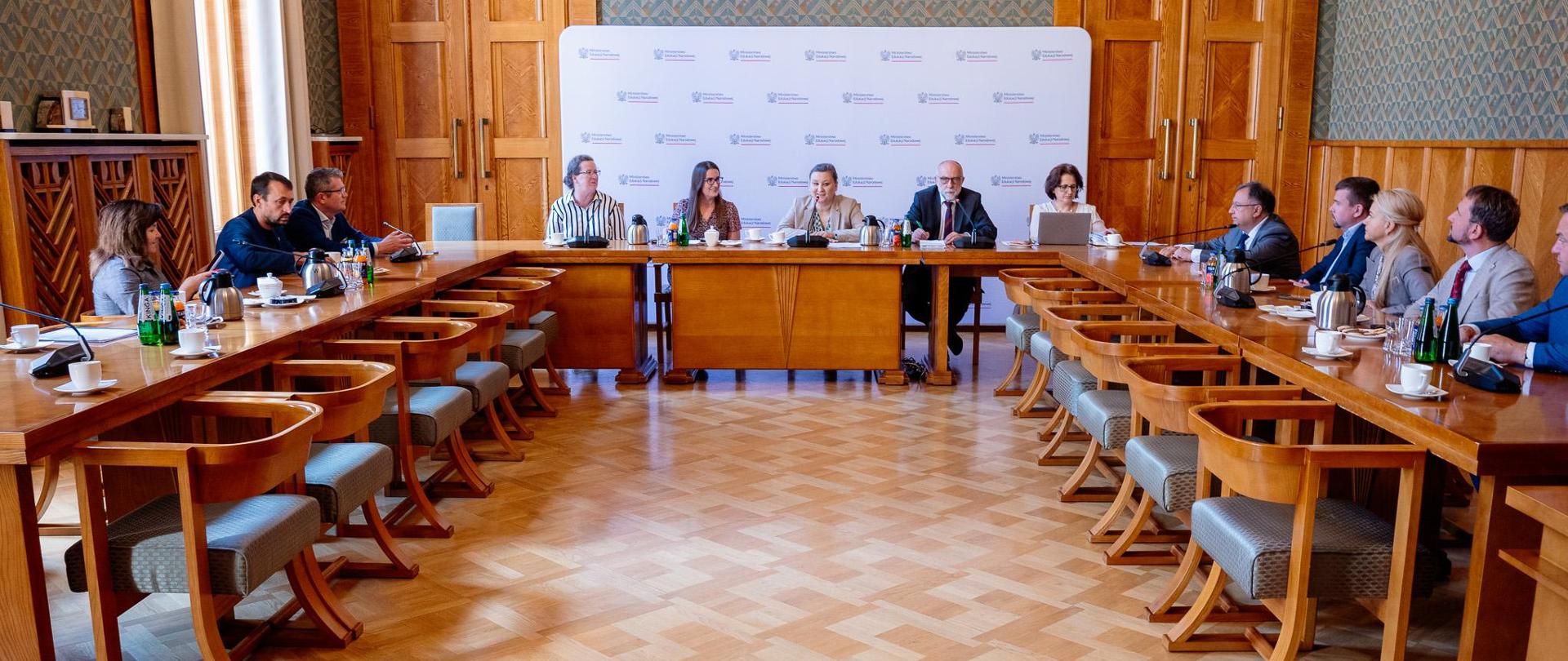 W sali stoją stoły i krzesła ustawione w formie odwróconej litery "u". Pośrodku siedzi wiceminister Paulina Piechna-Więckiewicz i zabiera głos. Za jej plecami stoi ścianka z logiem Ministerstwa Edukacji Narodowej. Przy pozostałych stołach siedzą uczestnicy spotkania. 