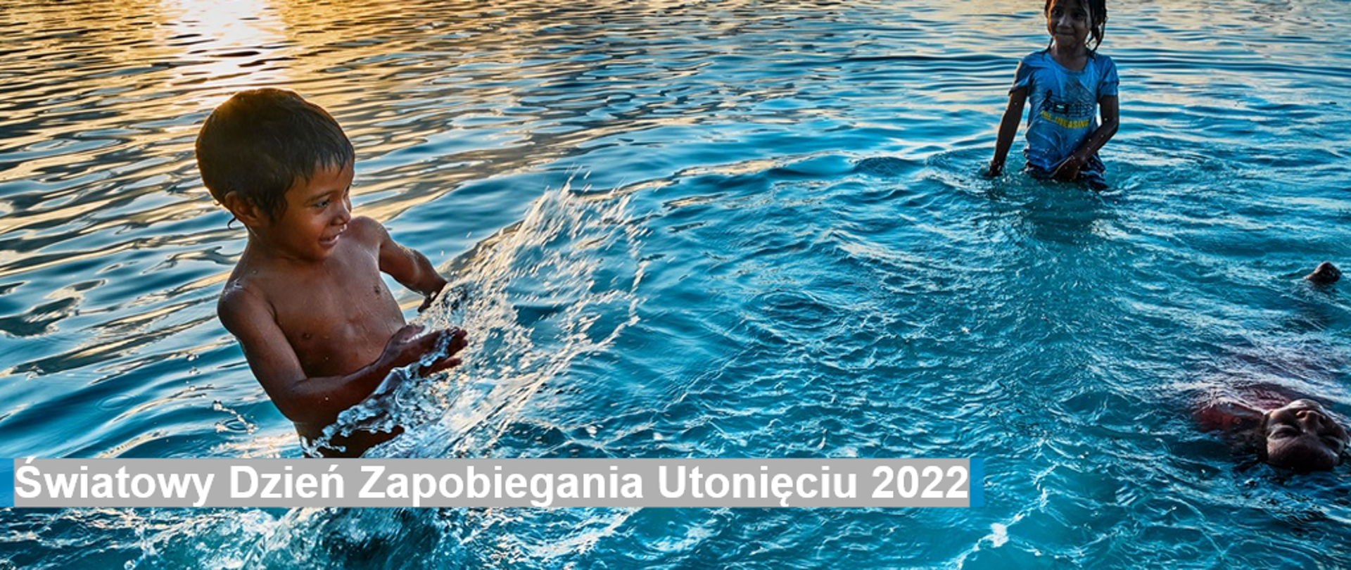 W wodzie kąpie się dwoje dzieci (dziewczynka i chłopiec) natomiast jeszcze jedna osoba płynie na plecach.