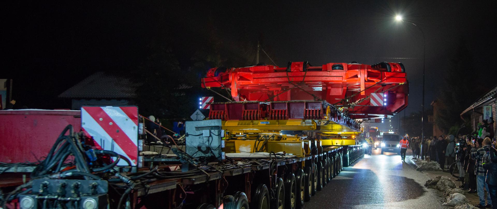 Na nocnej, mokrej od deszczu drodze jedzie ogromny, wieloosiowy transport specjalny. Na długiej platformie widać masywny, czerwony element konstrukcyjny oświetlony intensywnymi reflektorami. Po obu stronach ulicy stoją ludzie obserwujący przejazd. W tle jadą samochody techniczne, a całą scenę rozświetla latarnia uliczna oraz światła pojazdów.