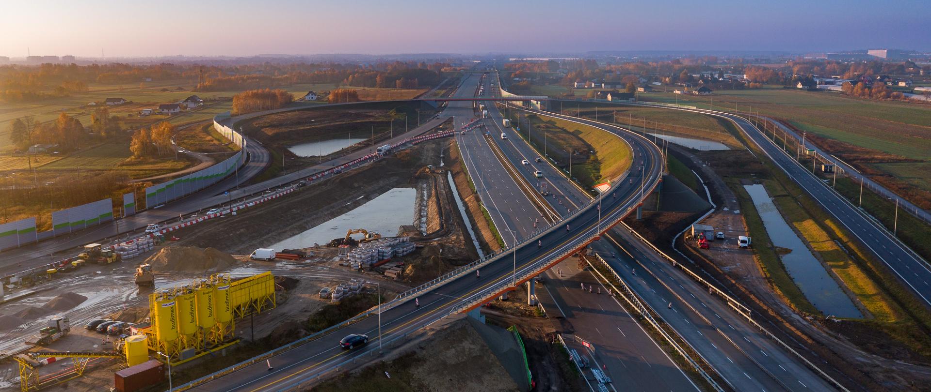 Widok na węzeł w porannym słońcu. Na pierwszym planie widać łącznicę węzła Warszawa - Katowice. Po prawej stronie zdjęcia nowa łącznica dla kierunku Łódź - Warszawa. W lewym górnym rogu fotografii zaplecze budowy z żółtymi silosami.