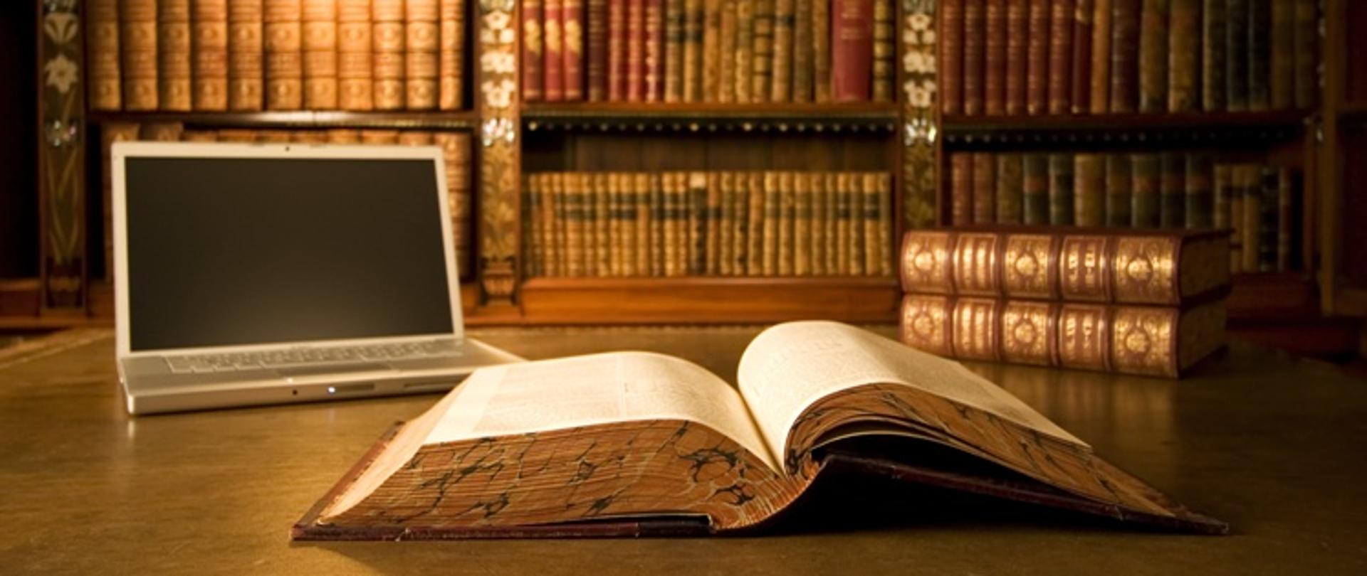 Na biurku leży otwarta duża księga i otwarty laptop, w tle widać półki pełne książek.