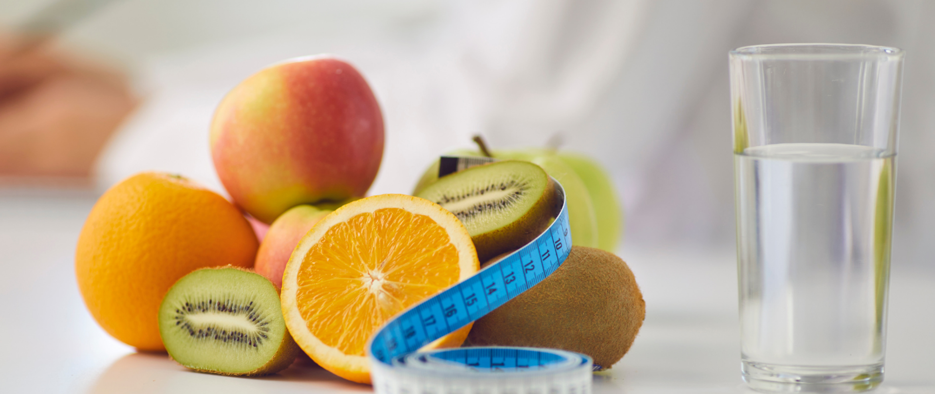 Na fotografii ukazano owoce: pomarańcze, jabłka, kiwi obok których leży miara. Z lewej strony owoców znajduje się szklanka wody