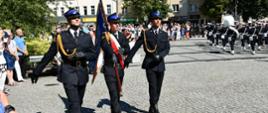 Zdjęcie przedstawia strażaków umundurowanych w mundury wyjściowe ze sznurami, maszerujących w trójce, tworząc poczet sztandarowy. Poczet maszeruje po placu brukowym, w tle widać zebranych uczestników obchodów. Strażak po środku niesie na ramieniu sztandar jednostki organizacyjnej PSP.