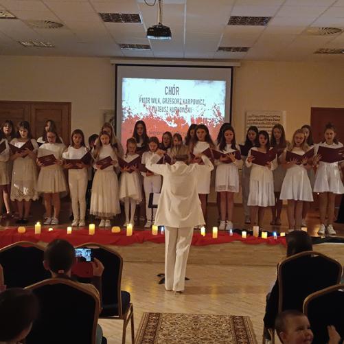 Zdjęcie przedstawia chór śpiewający pieśni patriotyczne. Chór jest ubrany na biało.W tle na ekranie widnieje napis chór, oraz skład zespołu nauczycieli akompaniujących: Piotr Wilk, Grzegorz Karpowicz, Mateusz Kurkowski. Ekran jest w kolorze biało-czerwonym. Dalej widzimy grafikę przedstawiającą partyturę, brązowe drzwi, oraz stół, zza którego wystaje biało-czerwona flaga. Przed chórem na podłodze leży czerwony materiał, a na nim białe zapalone świece. Przed sceną stoi Pani Ewelina Chrzanowska i dyryguje chórem. Po bokach widać publiczność słuchającą koncertu. 