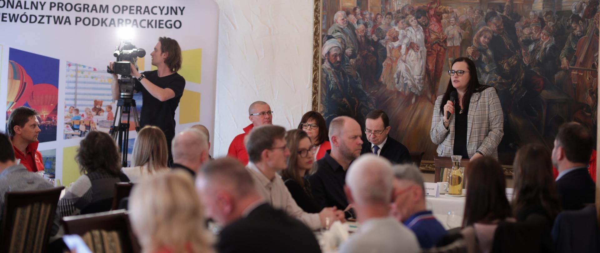Na sali przy stołach siedzi dużo osób. Na ostatnim planie na tle dużego obrazu stoi z mikrofonem minister Małgorzata Jarosińska-Jedynak.