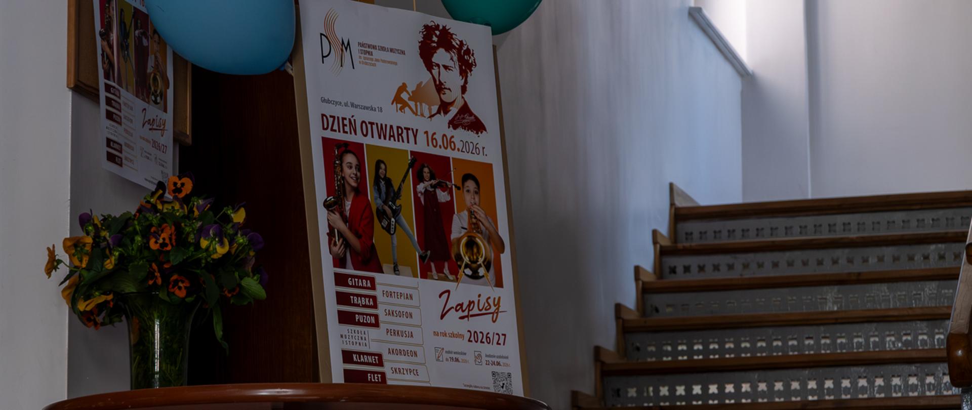 Korytarz szkoły z perspektywy wejścia głównego do budynku szkoły muzycznej. Stolik z kwiatami, za stolikiem sztaluga z plakatem informującym o dniu otwartym. Na górze plakatu doklejone baloniki z logo szkoły muzycznej.