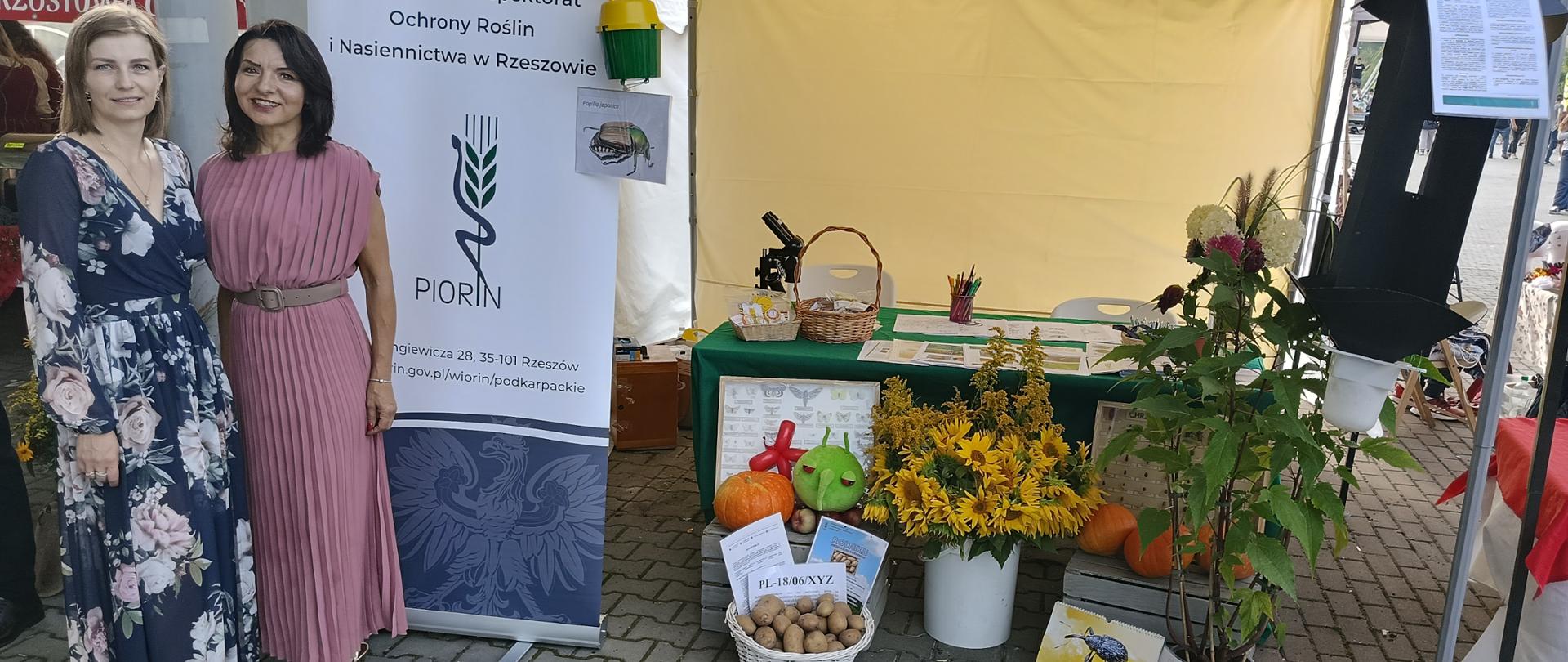 Dwie uśmiechnięte kobiety stoją przy banerze promocyjnym. Po prawej stronie stoisko z ulotkami informacyjnymi, gadżetami i mikroskopem. Z przodu dekoracje z kwiatów i warzyw.