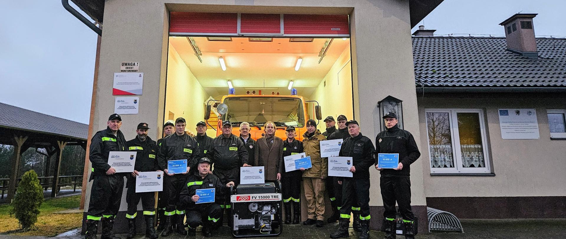 Wieczorowa pora. Przed oświetlonym garażem remizy stoją uczestniczy spotkania. Strażacy prezentują otrzymany sprzęt oraz dotacje. W tle wóz strażacki.