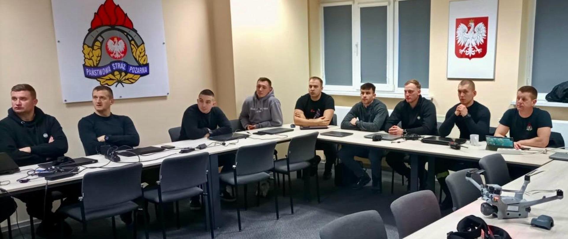 Sala szkoleniowa z grupą funkcjonariuszy siedzących przy stołach w kształcie litery U. Na stołach leżą kontrolery i sprzęt do obsługi dronów. Na ścianach widoczne są emblemat Państwowej Straży Pożarnej oraz godło Polski, co wskazuje na specjalistyczne szkolenie