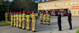 Na placu jednostki ratowniczo-gaśniczej, przed masztem, na którym powiewa flaga Rzeczypospolitej Polskiej, w dwóch szeregach naprzeciw siebie stoją strażacy w umundurowaniu bojowym. Przed masztem, twarzą do flagi, stoi trzech strażaków, którzy salutują. Na wprost masztu stoi również dwóch strażaków w umundurowaniu wyjściowym, którzy również salutują. W oddali widoczne samochody gaśnicze ustawione przed budynkiem remizy. 