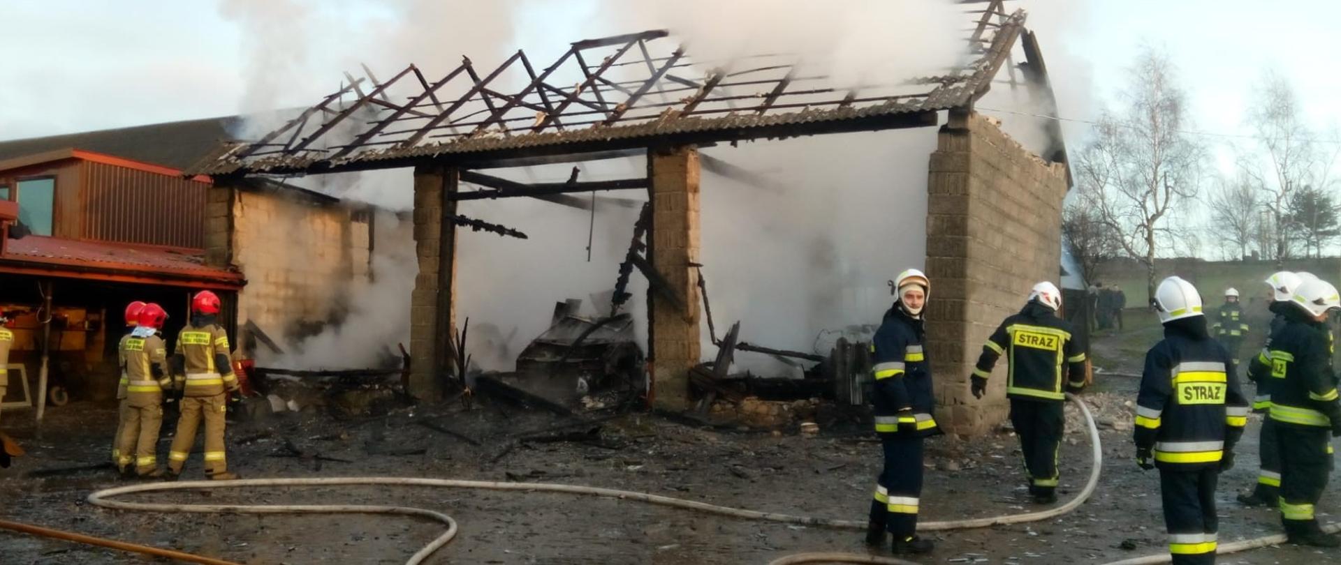 Zdjęcie przedstawia ruiny spalonej stodoły. Z lewej strony widać zabudowania gospodarcze, które udało się uratować. Na podwórzu stoją strażacy, a na ziemi leżą rozciągnięte węże strażackie. 