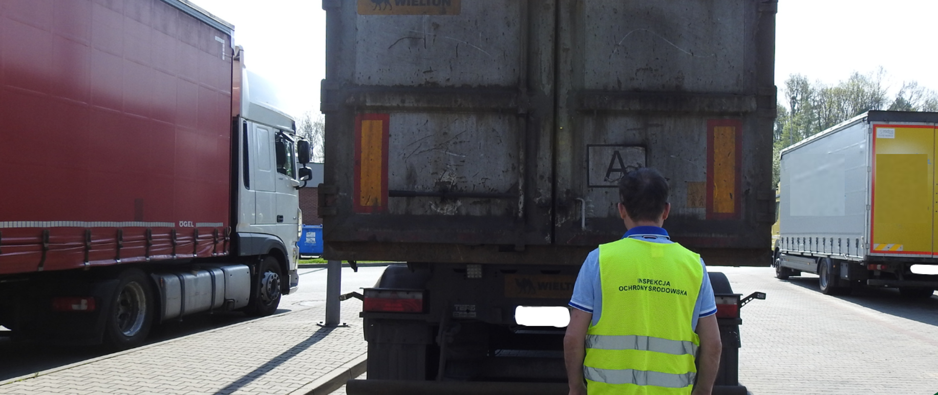 Mężczyzna obrócony plecami do zdjęcia z założoną kamizelką odblaskową, na której jest napis "Inspekcja Ochrony Środowiska". Przed mężczyzną na parkingu stoi samochód ciężarowy. Po boku widać inne samochody ciężarowe i motor.