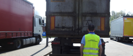 Mężczyzna obrócony plecami do zdjęcia z założoną kamizelką odblaskową, na której jest napis "Inspekcja Ochrony Środowiska". Przed mężczyzną na parkingu stoi samochód ciężarowy. Po boku widać inne samochody ciężarowe.