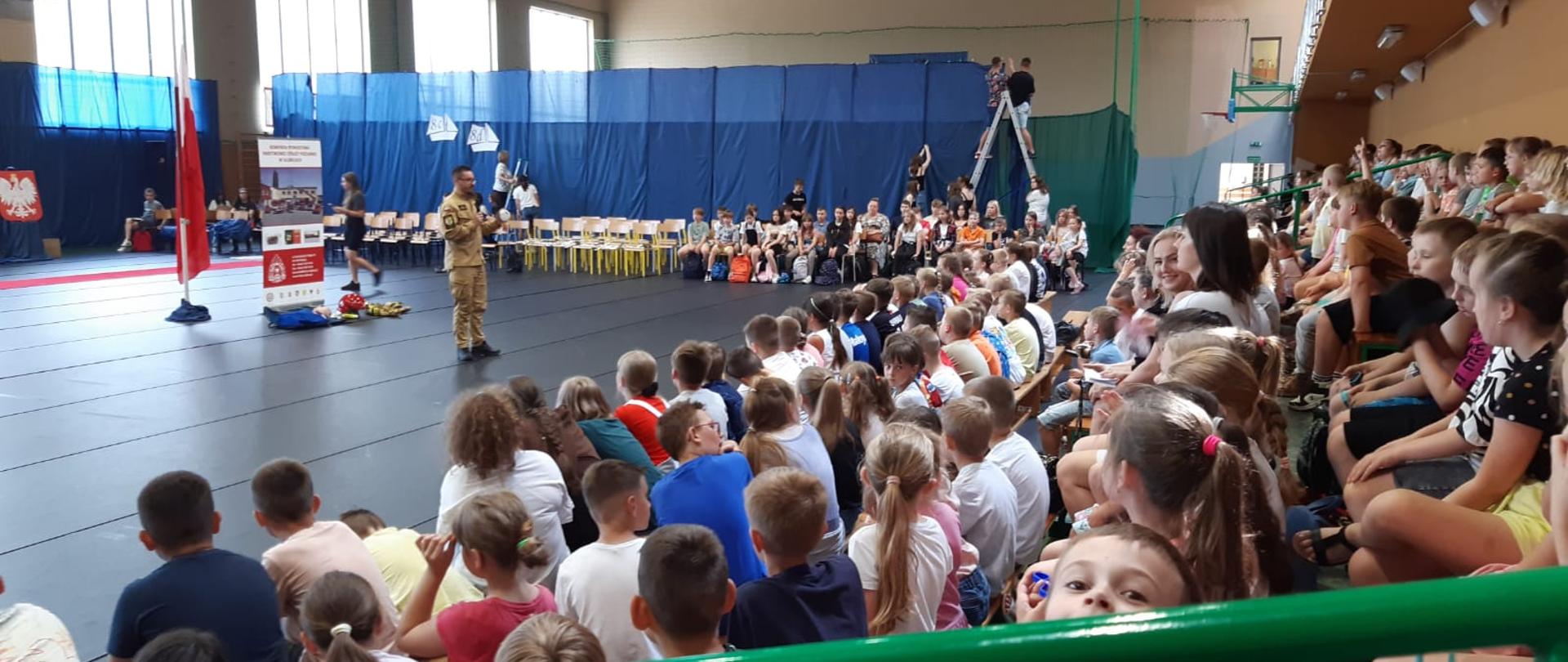 Wnętrze sali gimnastyczne w szkole. Na ławkach siedzi kilkadziesiąt dzieci. Na środku sali z mikrofonem umundurowany strażak PSP.