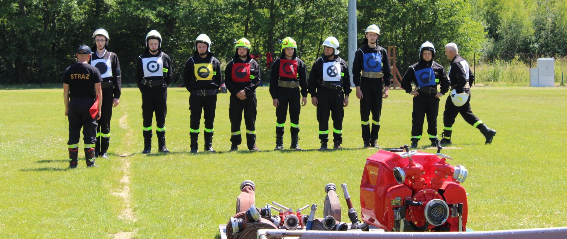 Na boisku w rzędzie stoją ustawieni strażacy gotowi do startu w ćwiczeniu bojowym. na pierwszym planie stoi motopompa