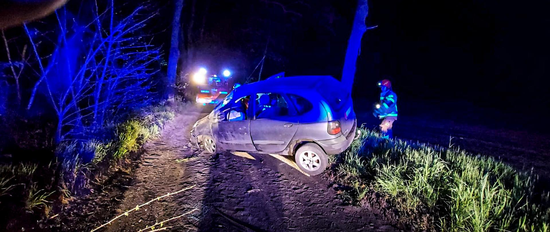 Zdarzenie miało miejsce w nocy. w środkowej części zdjęcia znajduje się samochód osobowy który stoi w poprzek drogi żużlowej przechylony na lewą stronę z rozbitym przodem, przebite przednie lewe koło. z tyłu za pojazdem jest strażak. w dalszej części zdjęcia jest samochód straży pożarnej oświetlający teren działań.