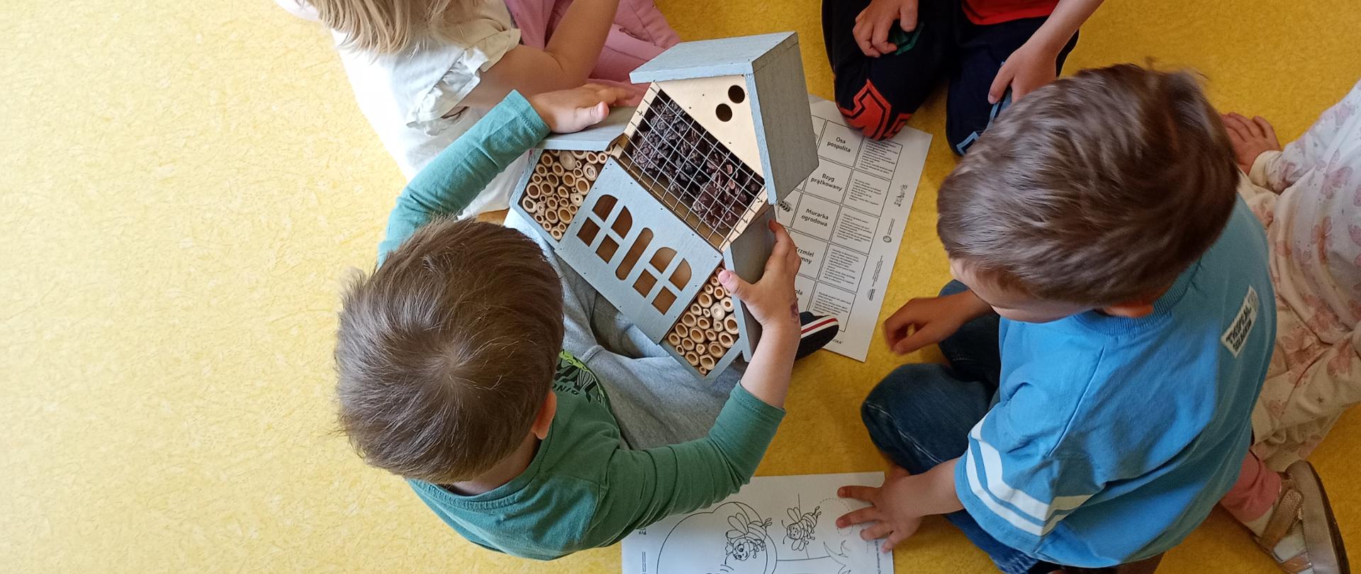 Dzieci siedzące na podłodze dotykające kartki z ilustracjami, domku drewnianego i nasion w woreczku.